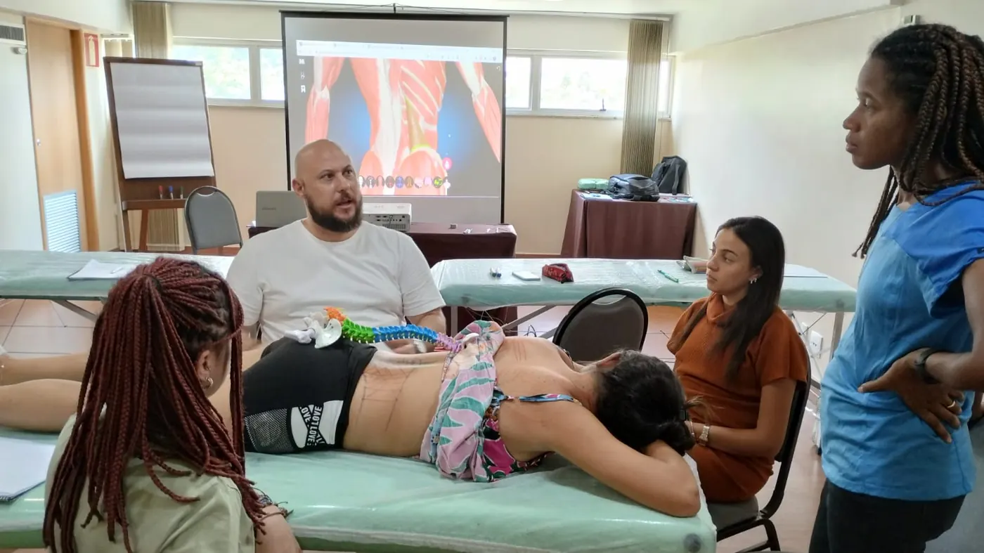Discussão de caso clínico com modelo da coluna vertebral e alunos acompanhando a avaliação na maca